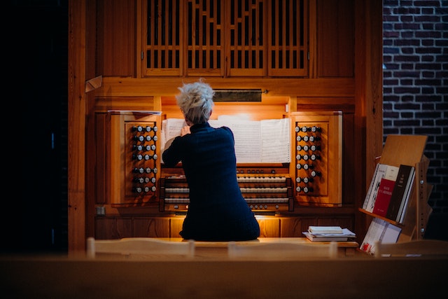 Das Bild zeigt eine Person, die an einer Orgel spielt. Sie sitzt mit dem Rücken zur Kamera und ist von hinten zu sehen. Die Orgel besitzt zahlreiche beleuchtete Registerknöpfe und Tasten. Auf der rechten Seite sind mehrere Notenbücher und Bücher auf einem Ständer zu erkennen.