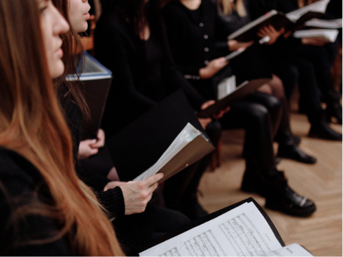 Das Bild zeigt eine Gruppe von Menschen, die in einer Reihe sitzen und Notenblätter in den Händen halten. Es scheint, als würden sie an einer Chorprobe oder einer musikalischen Aufführung teilnehmen. Sie tragen überwiegend schwarze Kleidung und halten schwarze Notenmappen, auf denen musikalische Notationen zu sehen sind.
